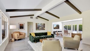 A spacious living room with a green sofa, beige armchairs, and a white rug. There are large windows and a view of greenery outside. - Sunset Ranch - Updated midcentury home with Pool & Spa! (Palm Springs)