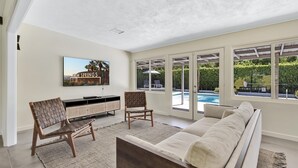 A spacious living room with a large window, a sofa, two chairs, a TV, and a pool view. - Sunset Ranch - Updated midcentury home with Pool & Spa! (Palm Springs)