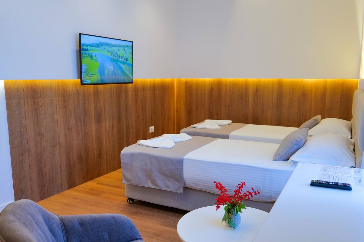 Una camera d'albergo moderna con una parete in legno decorativa, una televisione a schermo piatto sopra il letto, un piccolo tavolino con un vaso di fiori rossi e una poltrona.