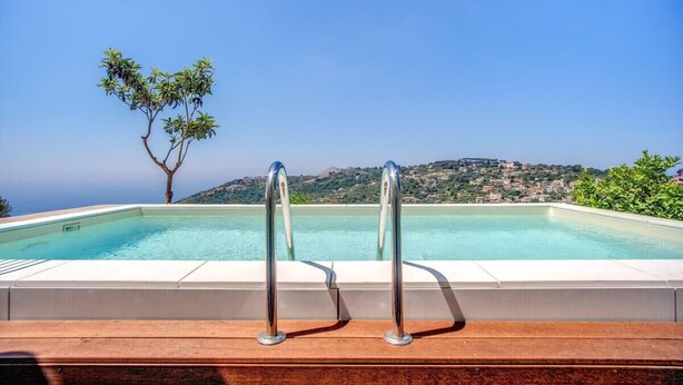 A swimming pool with metal railings, a wooden deck, and a view of a tree and distant buildings. - Villa La Luce: Town Centre, Sea Views, Pool & Concierge by Feeling Italy (Massa Lubrense)
