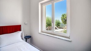 A bedroom with a bed, a red headboard, a small table, and a window with a view of greenery and a fence. - Villa La Luce: Town Centre, Sea Views, Pool & Concierge by Feeling Italy (Massa Lubrense)