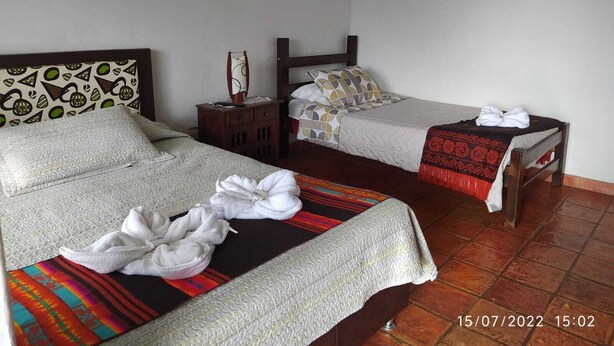 A hotel room with two beds, each with a patterned bedspread and pillows. There is a small wooden nightstand with a mirror and a lamp between the beds. The room has a tiled floor and plain walls. - Alquilo Hermosa Casa en Villa De Leyva (Villa de Leyva)