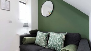 Ein Zimmer mit einem dunkelgrauen Sofa, zwei gemusterten Kissen, einem runden Spiegel an der Wand, einem kleinen Tisch mit einer Lampe und einem Fenster mit Jalousien.
