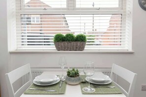 Mesa de comedor para dos personas con sillas blancas, una pequeña planta en maceta y dos copas de vino.