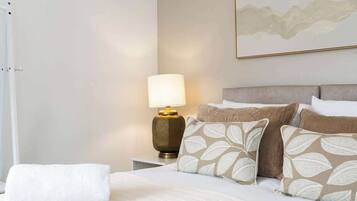 A neatly made bed with a white comforter and pillows, a bedside lamp, and a framed artwork on the wall.