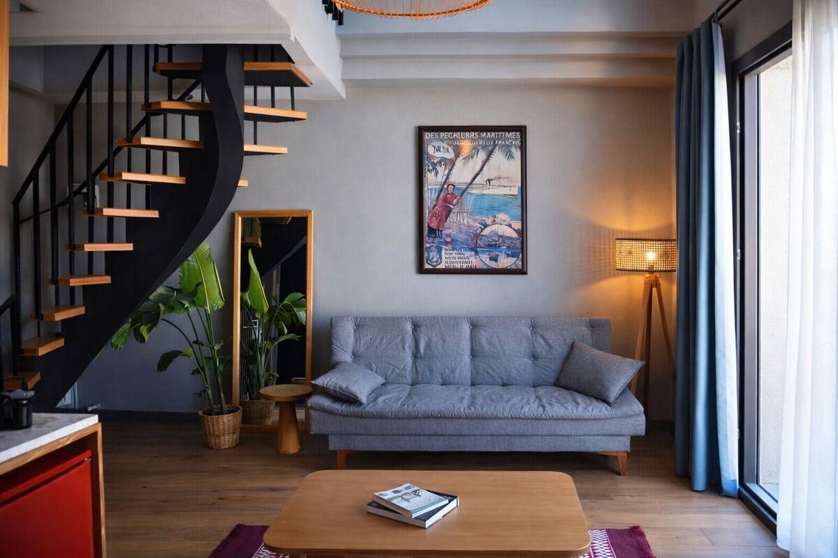 A modern living room with a grey sofa, wooden staircase, and a large window.
