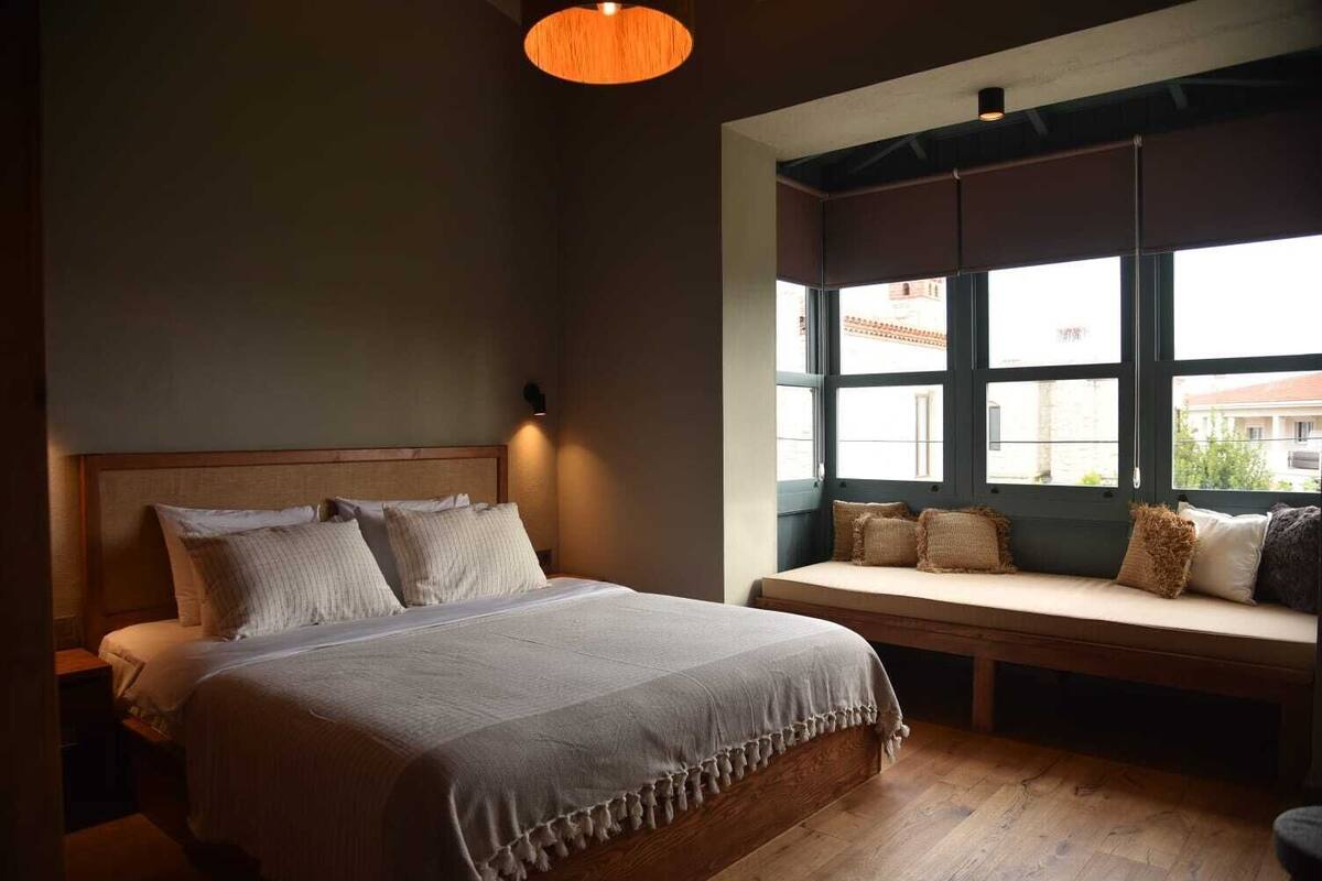 A bedroom with a large bed, a bench with cushions, a wall-mounted light, and a window with a view of buildings.
