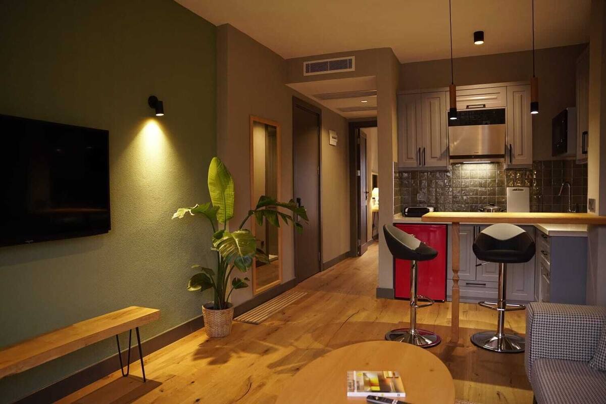 A modern living room with a wooden bench, a round coffee table, and a potted plant. A kitchen area with a red bar stool and a dining table is visible in the background.