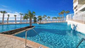 En swimmingpool med kristallklart blått vatten, omgiven av palmer och en vit byggnad med balkonger.