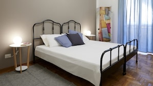 Una cama bien tendida con colchón blanco y estructura de metal negro, dos mesitas de noche con lámparas, una alfombra gris y un cuadro abstracto grande en la pared.