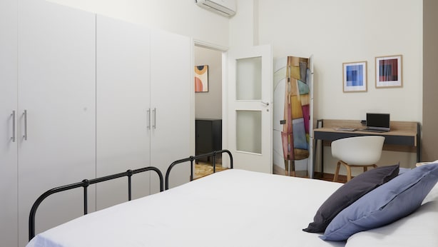 Un dormitorio con una cama, un escritorio y una silla. Hay un armario y una puerta que da a otra habitación.