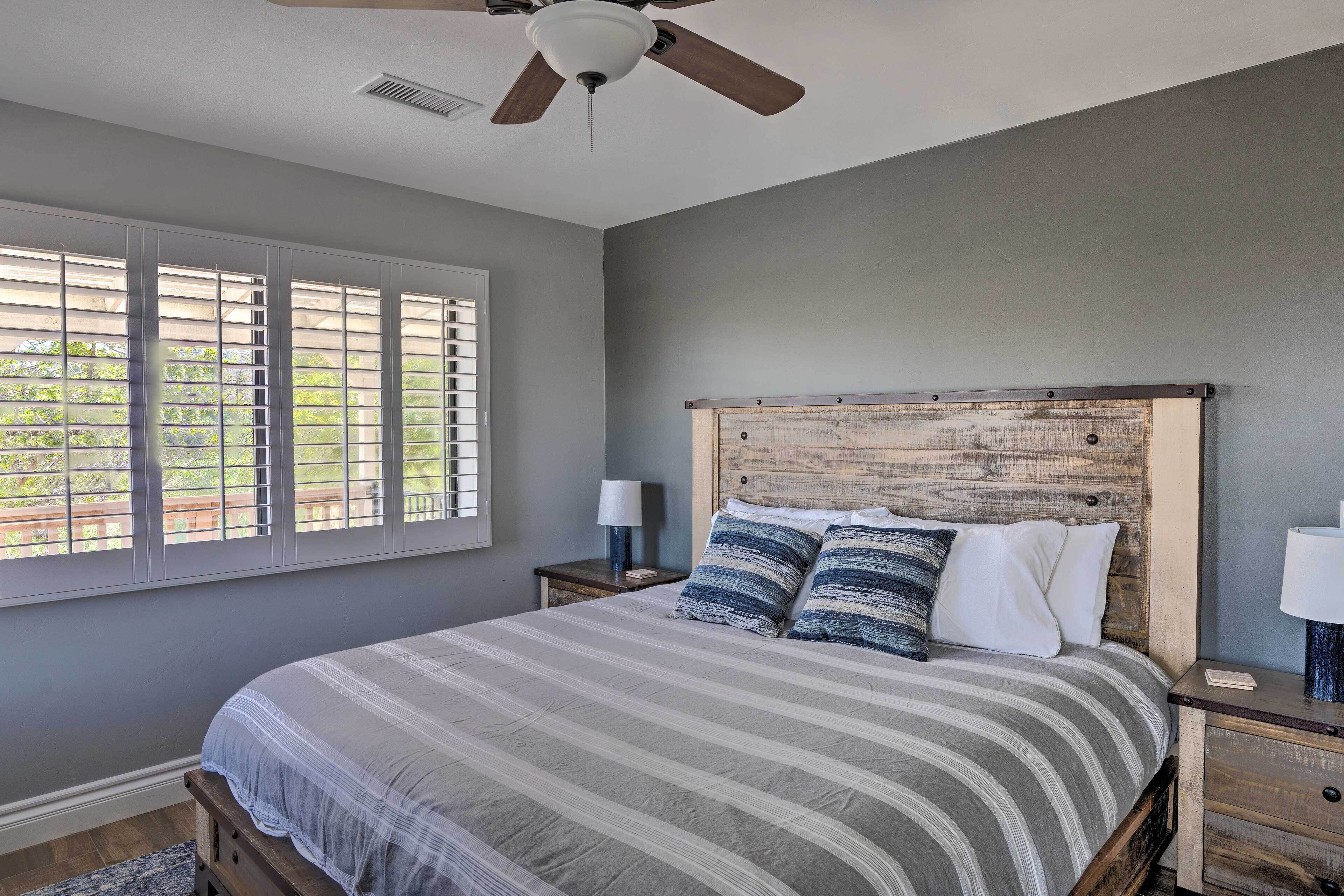 Une chambre à coucher avec un grand lit, un ventilateur de plafond, une fenêtre avec des stores et une table de chevet avec une lampe.