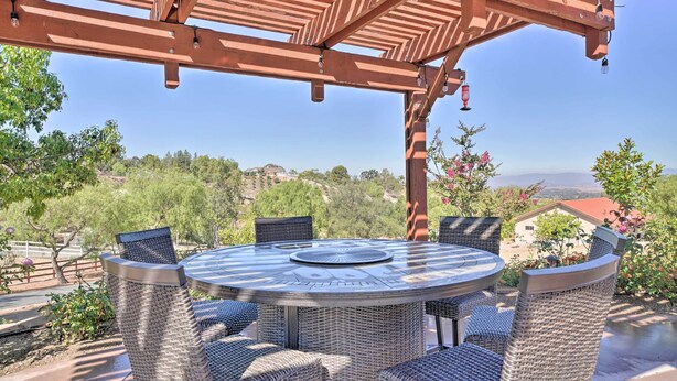 A round table with wicker chairs under a wooden pergola, overlooking a scenic landscape with trees and a distant view of a house.