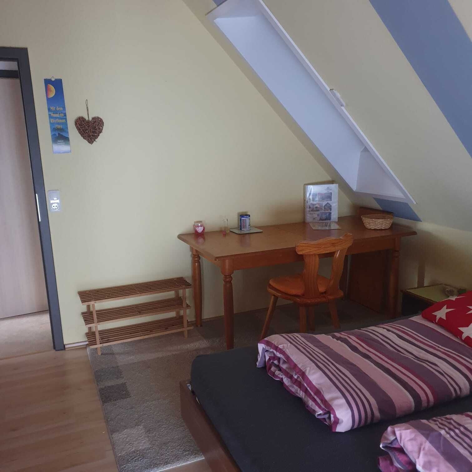 A small, cozy room with a sloped ceiling, a wooden desk, a chair, a bed with striped bedding, and a small shelf with items.