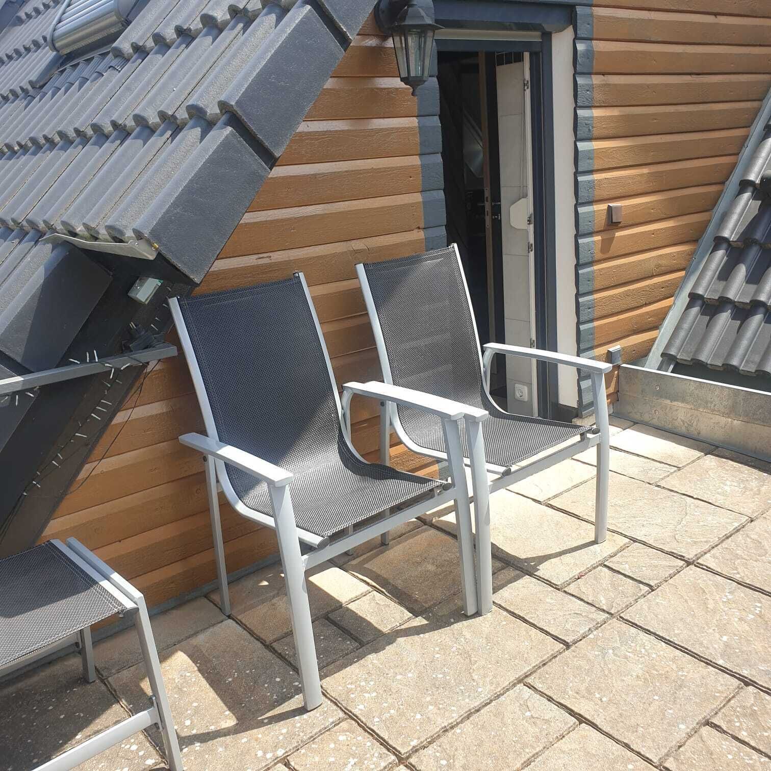 Dos sillas de metal con asientos de malla y estructura blanca, ubicadas en un patio de baldosas de piedra junto a un edificio de madera con techo inclinado.