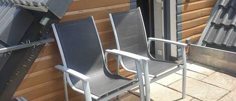 Dos sillas de metal con asientos de malla y estructura blanca, ubicadas en un patio de baldosas de piedra junto a un edificio de madera con techo inclinado.