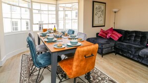 A dining area with a wooden table set for four, surrounded by blue chairs. There is a dark-colored sofa with a red pillow in the adjacent living area.