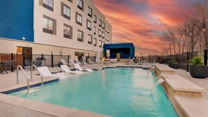 Une piscine avec des chaises longues, un bâtiment doté de nombreuses fenêtres et un ciel au coucher du soleil.