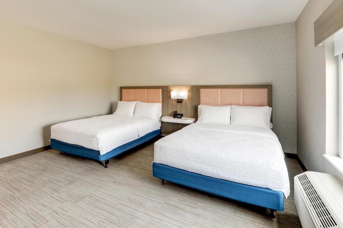 A hotel room with two beds, a nightstand, a lamp, and a window.