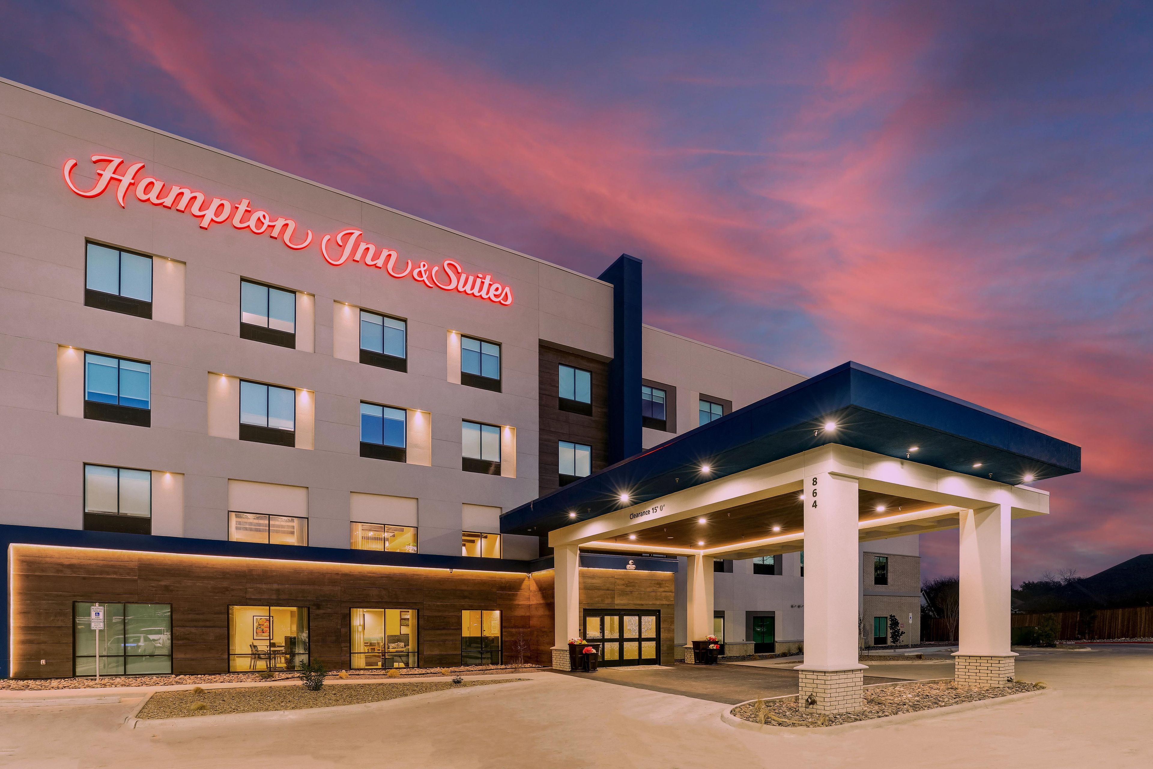 A Hampton Inn & Suites hotel with a modern facade, a covered outdoor area, and a clear sky.