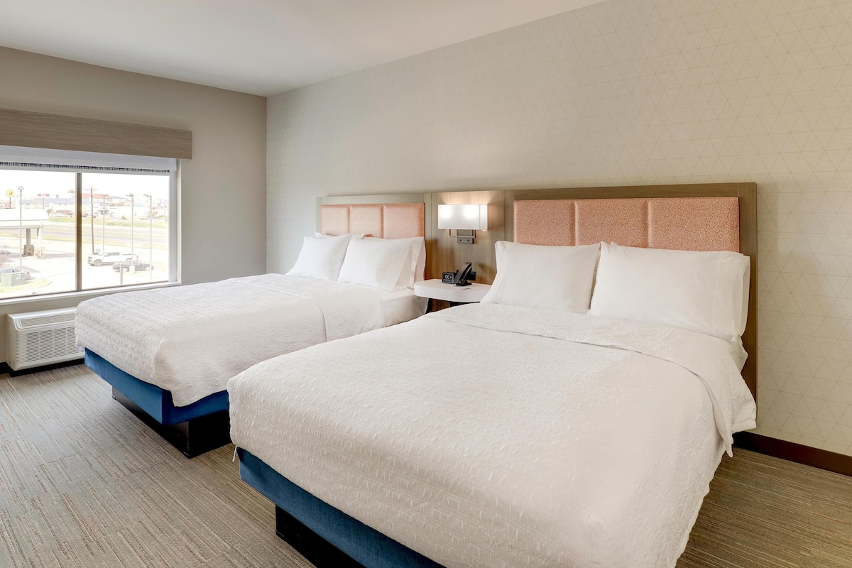 A hotel room with two beds, a window with a view, a bedside table with a lamp, and a wall-mounted air conditioning unit.