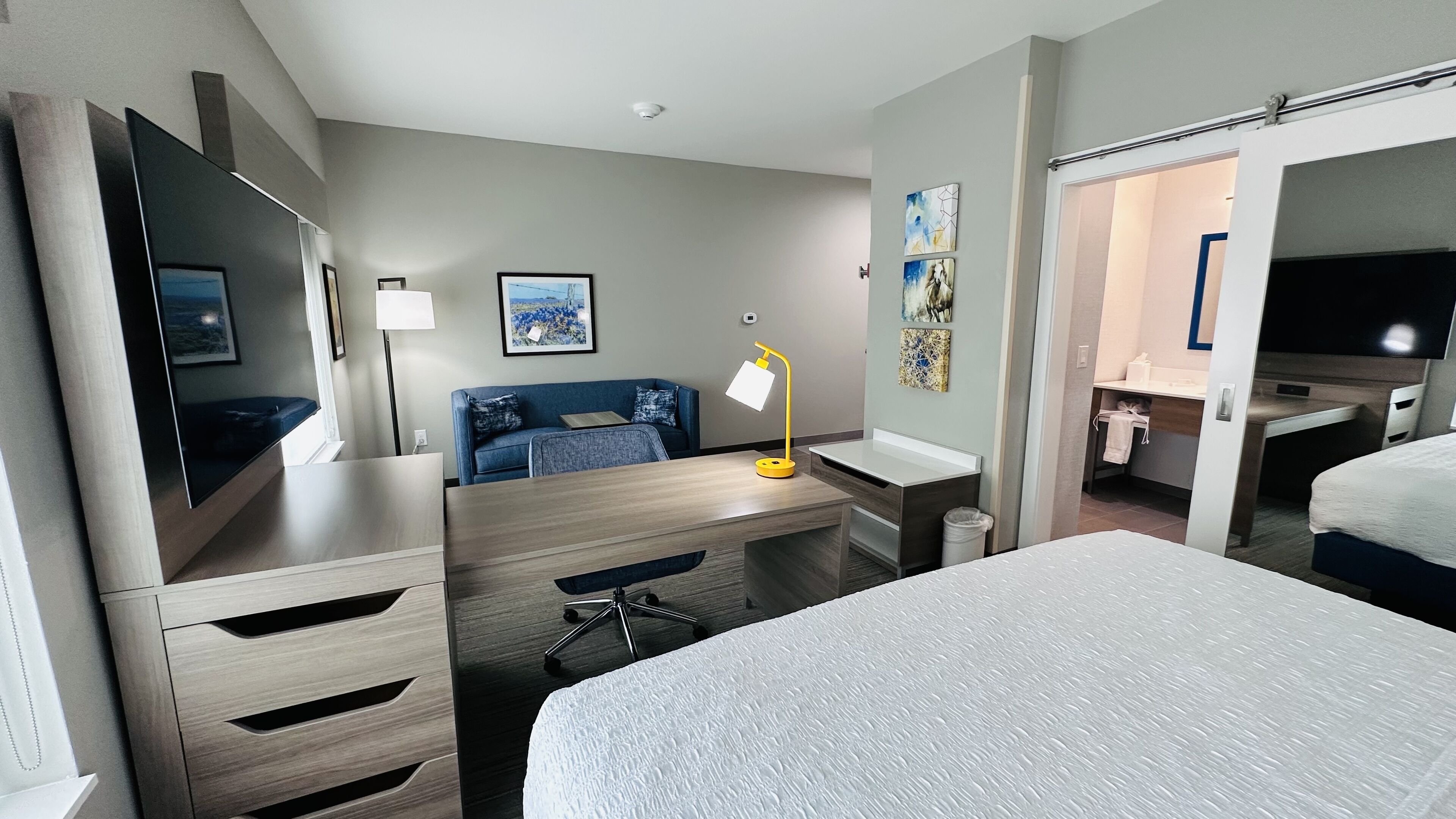 A hotel room with a bed, a desk with a lamp, a TV, a sofa, and artwork on the walls.