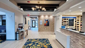 Un hall d’hôtel comprenant un comptoir de réception, un espace où l’on peut se servir du café, un tableau accroché au mur, et un panneau indiquant l’emplacement de la salle de petit-déjeuner et des salles de réunion.