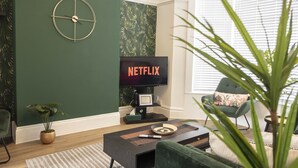 A living room with a green wall, a round clock, a flat-screen TV displaying 'NETFLIX', a coffee table, a green armchair, and a potted plant. - The Callie by Sasco Apartments (Blackpool)