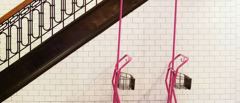 A pink hand truck with a basket against a white brick wall and a black metal railing.