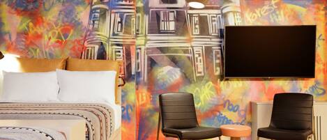 Habitación de hotel con cama, dos sillas negras, una mesita redonda, televisor y un mural colorido en la pared.