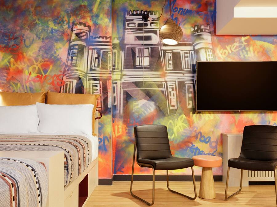 Hotellihuoneessa on sänky, kaksi mustaa tuolia, pieni pyöreä pöytä, televisio ja värikäs seinämaalaus.