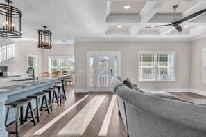 A modern kitchen with a bar area, wooden flooring, and a gray sofa in the living area. - Carolina Sunshine: Spacious Home Less Than Half A Mile to Heron Lookout Park and (Oak Island)