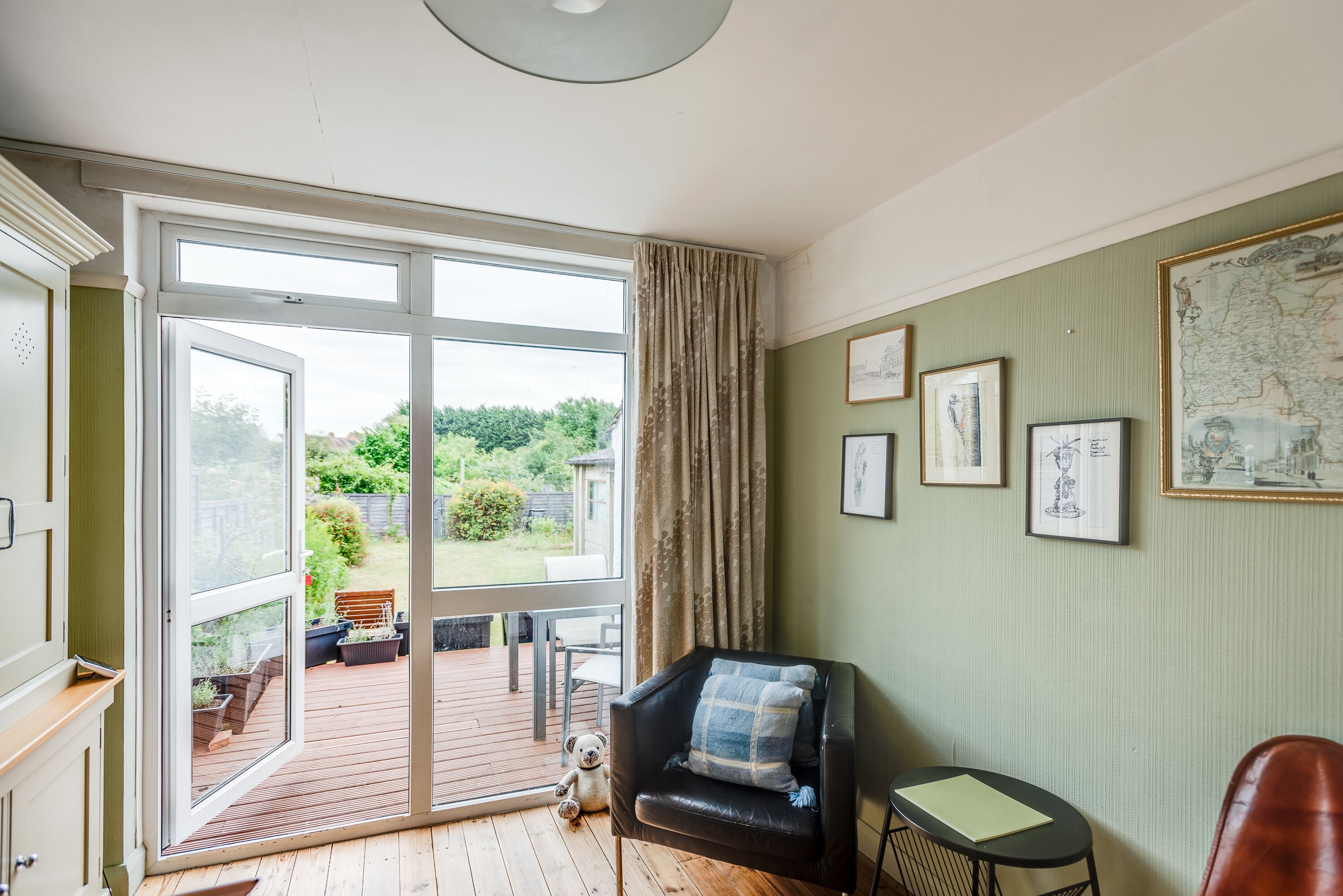 Une pièce dotée d’une grande porte-fenêtre coulissante donnant sur une terrasse, d’un fauteuil en cuir noir avec un coussin à carreaux, d’une petite table sur laquelle se trouve un dossier vert, et d’œuvres d’art encadrées accrochées au mur.
