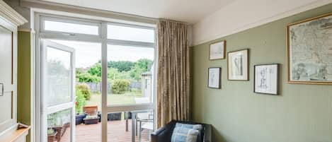 A room with a large sliding glass door leading to an outdoor deck, a black leather armchair with a plaid cushion, a small table with a green folder, and framed artwork on the wall.