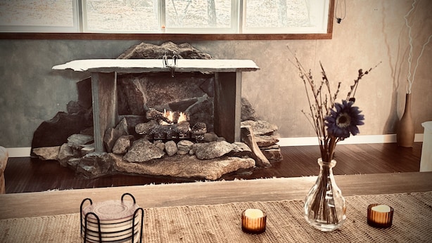 Ein Kamin mit Holzscheiten, eine Vase mit Blumen und Kerzen auf einer gewebten Matte.