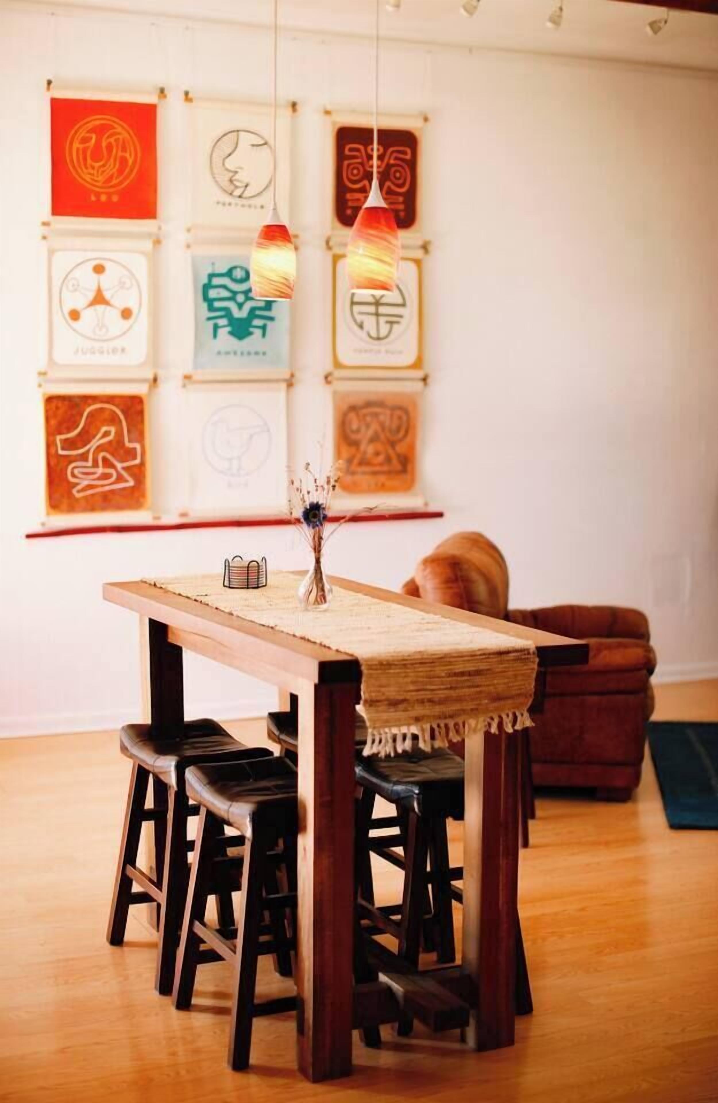 Un comedor con una mesa de madera y cuatro taburetes, un jarrón con flores secas y una pared decorada con obras de arte enmarcadas.