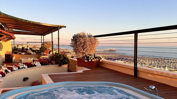 A hot tub on a wooden deck with a view of the sea and a building with outdoor seating. - Penthouse Piccadilly with Jacuzzi (Lido di Camaiore)