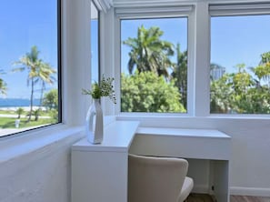 A modern room with a white desk, a vase with greenery, and a view of palm trees and the ocean. - Miami Villa: Resort Style Oasis with Pool, Hot Tub, Game Room / Sleeps 16+ (Miami Shores)