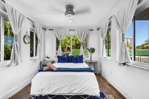 Ein Schlafzimmer mit Bett, Vorhängen, einem Nachttisch, einem kleinen Tisch, einem Deckenventilator und Blick auf Grünfläche.