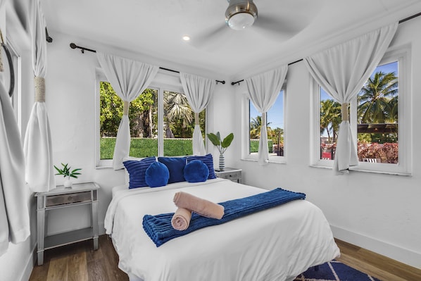 A bedroom with a bed, white curtains, a nightstand, a plant, and a view of greenery.