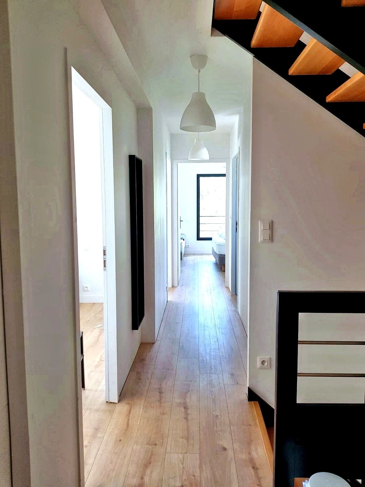 A long, narrow hallway with wooden flooring, white walls, and a white pendant light. There is a door on the left and a staircase on the right.