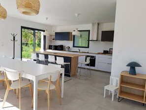 A modern kitchen with white cabinetry, a dining area with a white table and chairs, and a large window providing a view of greenery outside.