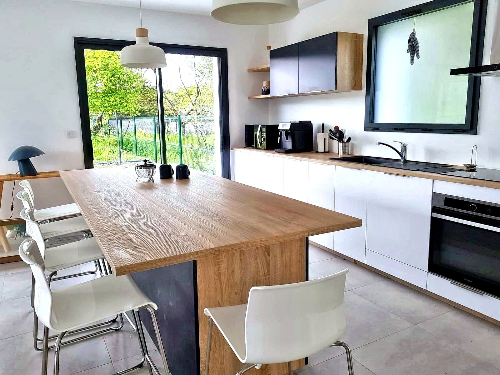 A modern kitchen with a wooden island, white chairs, a black countertop, and a large window.