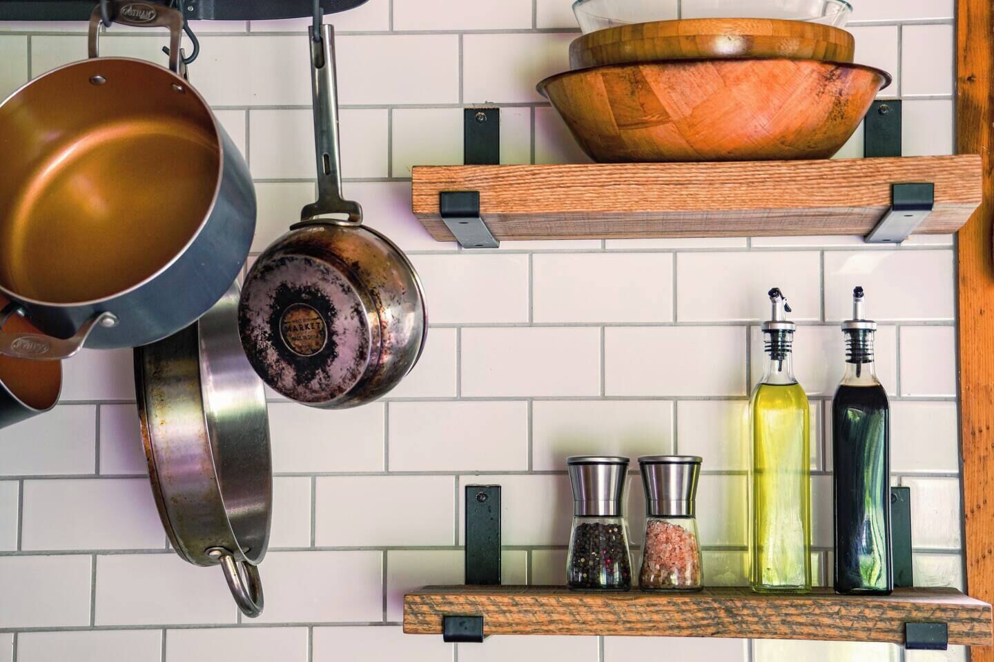 A kitchen with hanging pots, a wooden bowl on a shelf, and bottles of oil and spices.