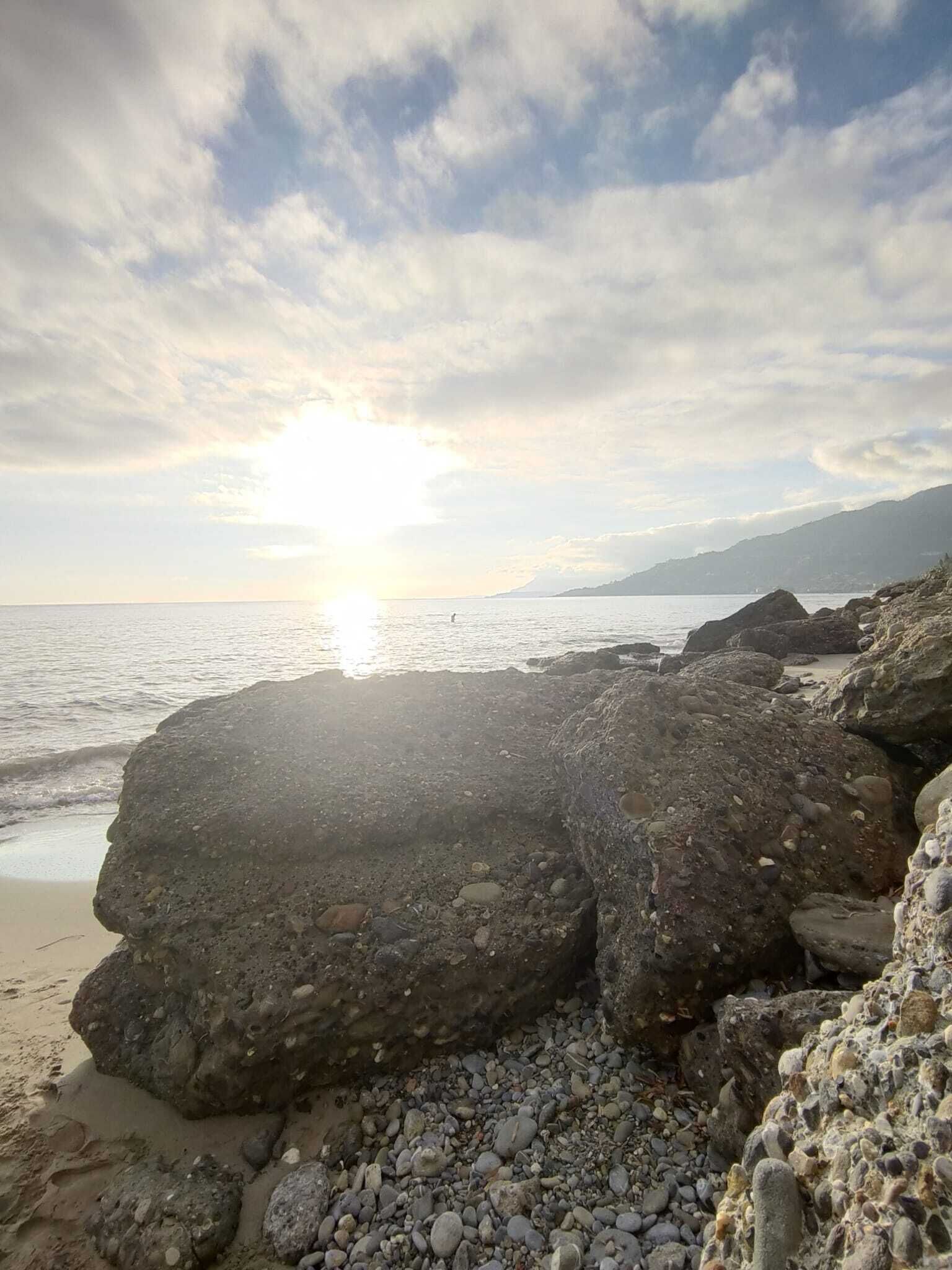Una playa rocosa con grandes rocas y piedras, un mar tranquilo y una puesta de sol al fondo.