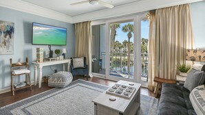A living room with a flat-screen TV, a coffee table, a sofa, a side table, a bookshelf, and a sliding glass door leading to a balcony with palm trees. - 97 Steps to Beach,  Beautiful Condo, Balcony Sleeps 5 (Santa Rosa Beach)