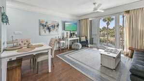 A spacious living room with a dining area, a flat-screen TV, and a large window with a view of palm trees. - 97 Steps to Beach,  Beautiful Condo, Balcony Sleeps 5 (Santa Rosa Beach)