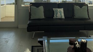 Ein Wohnzimmer mit einem dunklen Sofa, zwei Kissen und einem Glastisch. Große Fenster bieten Blick auf den Ozean und Palmen.