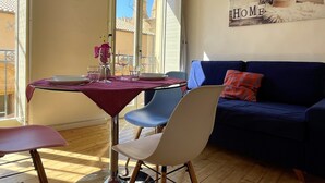 Un coin repas avec une table dressée pour deux personnes, un canapé bleu et une vue sur les bâtiments par la fenêtre.
