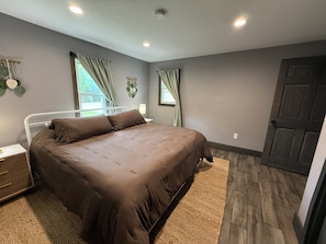 A bedroom with a bed, nightstands, a window with curtains, a door, and a wall-mounted light fixture.